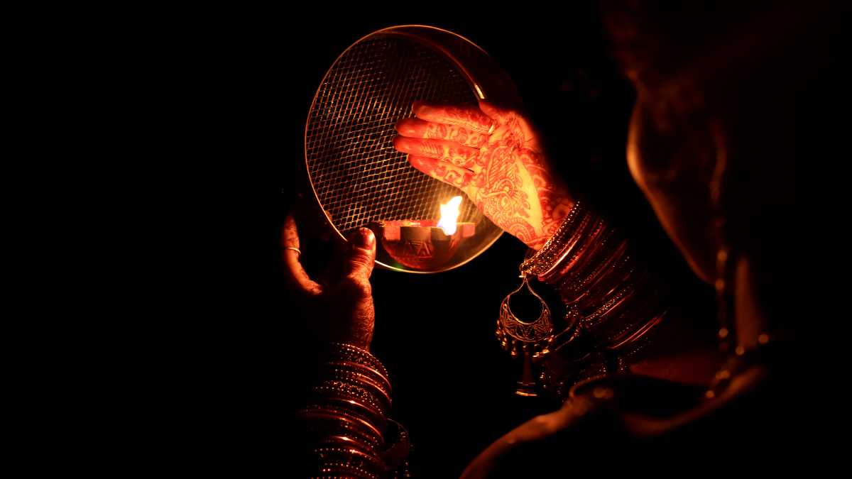 Hands covered in henna and glass bangles holding a sieve above a lit diya in the dark - Divine Sansar UAE
