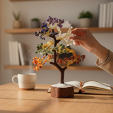 Seven Chakra Crystal Tree of Life self-charging on a wooden base, held by a hand on a table with a cup and book in the background.