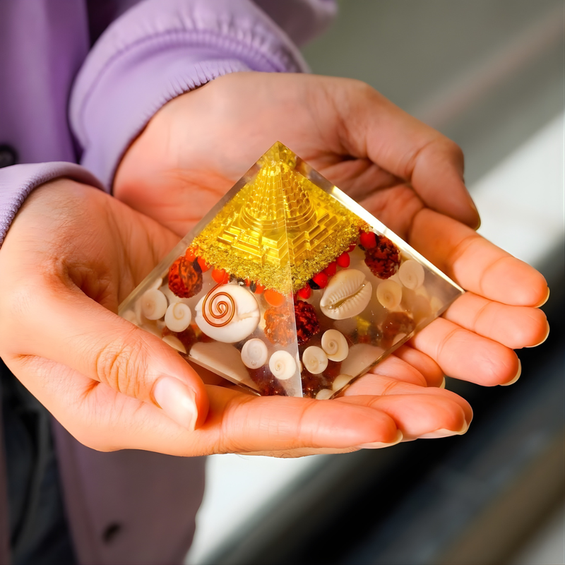 Divine Sansar -Orgonite Shree Yantra Prosperity Pyramid containing a golden Shri Yantra and natural shells is held gently in two hands. 