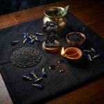 Divine Sansar-Ceremonial setup with a Shani dev statue, bowls of seeds, and a lit candle on a dark cloth.