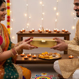 Couple exchanging a gold Diwali return gift box at Lakshmi Puja with diyas and rangoli - Divine Sansar